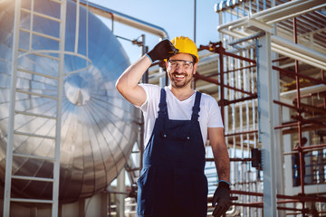 Happy caucasian worker in overall and with helmet on head posing in front of oil storage tank.