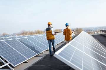 View on the rooftop solar power plant with two engineers walking and examining photovoltaic panels....