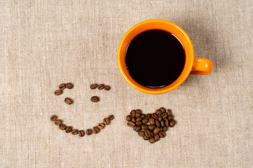 A mug of hot black coffee, coffee beans in the form of a smile and a heart on a fabric background.