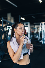 Young attractive woman after successful workout in modern fitness gym holding glass of protein...