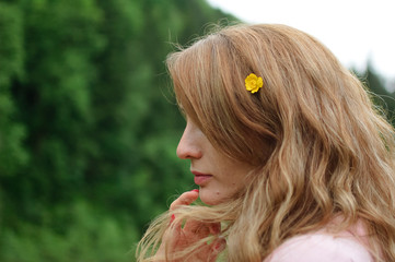 Outdoors portrait of young calm blonde woman in pink clothes with little yellow flower in her long hair on the hill with green forest background during spring or early autumn in the mountains