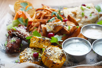 a plate of beer snacks. Corn with cheese, smoked cheese, chicken wings, potato slices with cheese, envelopes with meat vegetables, herbs and three sauces.