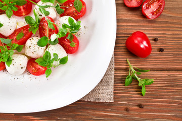 Caprese salad with ripe red cherry tomatoes, mozzarella cheese balls, young basil leaves, spices with sprouts of pea on a wooden background.