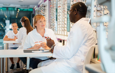 Professional African-American ophthalmologist helping mature woman to choose glasses in optical store