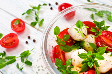 Caprese salad. Ripe red cherry tomatoes, mozzarella cheese balls, young basil leaves and spices with young sprouts of pea on a white wooden background.