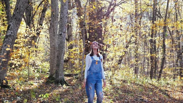 Happy young woman having fun, laughing and throwing yellow leaves at autumn forest in sunny day. Leaves is falling in slow motion.