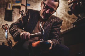 Focused mature cobbler is fixing sole for boots using hammer.