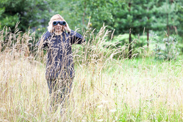 Mature woman an ornithologist hidden in high grass watching with binoculars in nature, copyspace