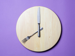 Intermittent fasting and skip breakfast concept - empty wooden round tray or trencher with cutlery as clock hands on lilac background. Eight hour feeding window concept or breakfast time concept
