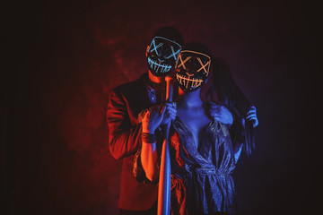 Man and woman in masks are posing for photographer with baseball bat in red and blue lights.