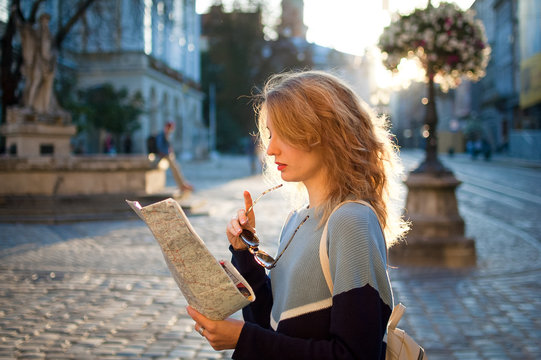 Side View Of Young Woman In Casual Sweater Is Looking For Direction Using Paper Map Early In The Morning In Ancient European City On Empty Square