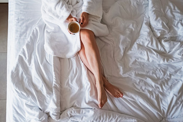 Fototapeta na wymiar Unrecognizable woman in white bath robe enjoying morning coffee while sitting on her unmade bed.