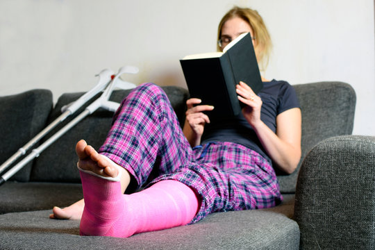 Woman with plaster cast sitting on sofa and read a book. She has lifted her leg to couch. Copy space.