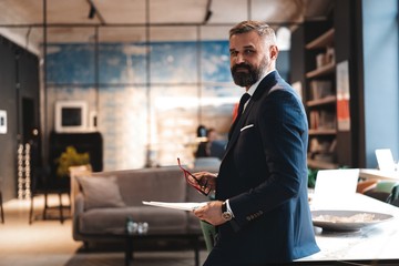 Stylish businessman standing near table in the office and reading documents