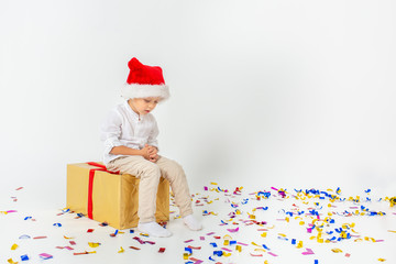 Little sad boy in santa helper hat sitting on a giftbox with red ribbon, white isolated background, confetti. Christmas, winter, new year concept.