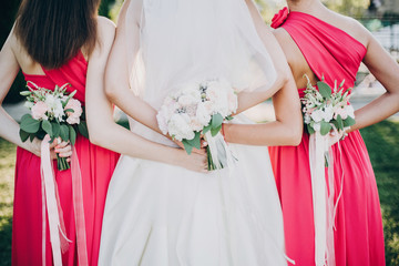 Gorgeous bride posing with bridesmaids in pink dresses, holding stylish bouquets on back in warm...