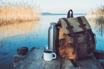 Fototapeta Enameled mug of coffee or tea, backpack of traveller and thermos on wooden pier on tranquil lake. obraz