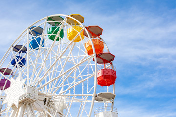 Ferris wheel with white structure and colored cabins on background of blue sky
