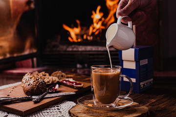 Pouring milk from porcelain milk jug into cup filled with tea - white tablecloth and fire...