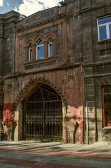 Narrow two storey house of red tuff with oval Windows and an entrance from an arch covered with carved ornaments with forged black gates