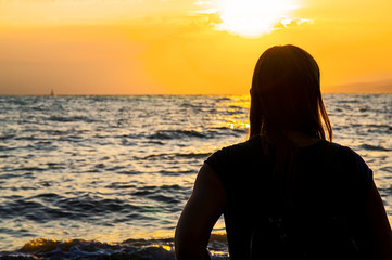 The girl on the seashore looks at the orange sunset.