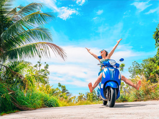 Crazy funny woman with flying hair riding a motorbike on a blue sky and green tropics background. Young bizarre girl with dark hair in sunglasses on a blue scooter in vintage style racing downhill
