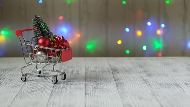 Hand puts mini shopping cart with christmas tree and gifts on the background of led lamps garland. Concept of Christmas sale