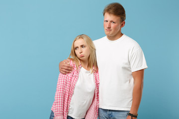 Young fun couple two friends guy girl in white pink empty blank design t-shirts posing isolated on pastel blue background studio portrait. People lifestyle concept. Mock up copy space. Looking upset.
