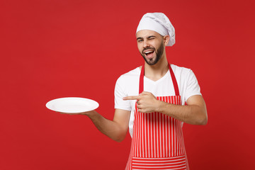 Cheerful chef cook or baker man in striped apron toque chefs hat isolated on red background. Cooking food concept. Mock up copy space. Pointing index finger on empty blank plate with place for food.