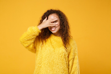 Smiling young african american girl in fur sweater posing isolated on yellow orange wall background, studio portrait. People lifestyle concept. Mock up copy space. Covering face with hand, peeping.