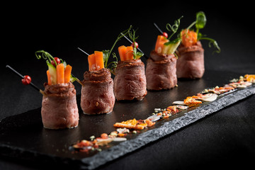 Mini roast beef rolls with vegetables, on a black slate plate, on a black background. Fusion food concept, low key, copy space.