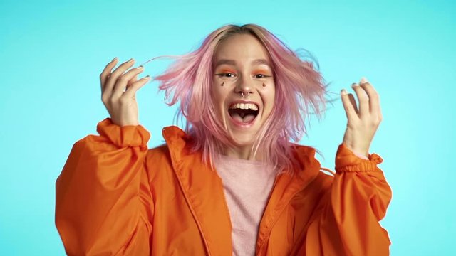 Excited happy screaming woman with unusual pink hairstyle. Portrait of young girl with ecstatic face expressions. Mixed race female shocked model on blue background. 