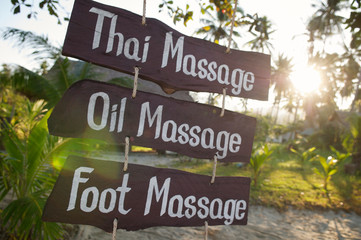 Rustic wooden sign offering different massages (Thai, Oil and Foot) hanging at a tropical beach in Thailand at sunset