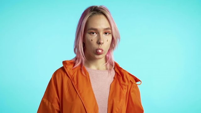 Playful girl with pink hair blows bubblegum, chewing gum in slow motion. Hipster woman with colorful dyed hair, piercing stands on blue background.