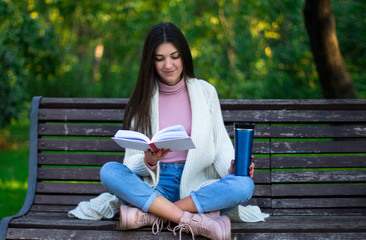 young female is reading a book on the bench in the park, thermo mug on the bench