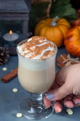 Woman's hand take a glass with hot white chocolate with pumpkin and spices, garnished with whipping cream and cinnamon, autumn decorations on background, vertical