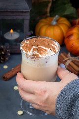 Woman's hand take a glass with hot white chocolate with pumpkin and spices, garnished with whipping cream and cinnamon, autumn decorations on a background, vertical