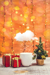 White mug with snowing cloud, present boxes and little Christmas tree with bokeh lights on a wooden table