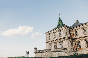 Gorgeous wedding couple walking in sunlight near old castle in beautiful park. Stylish beautiful bride and groom posing on background of ancient building and nature