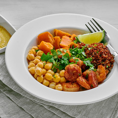 Grilled shrimps and sweet potato with black quinoa, chickpeas in a soup plate with humus and a glass of water on a white table.