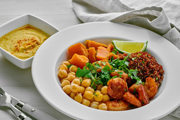 Buddha nourish bowl with garlic grilled shrimps, baked sweet potato, black quinoa, chickpeas, humus and lime on a table.
