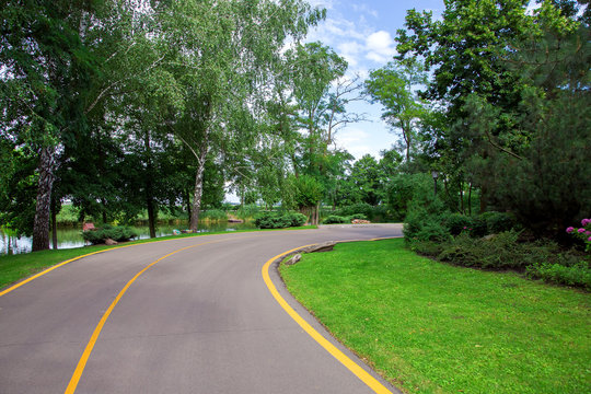 asphalt road with yellow markings leaving for a turn in the park with a green lawn and trees on the side of the road and a river with a blue sky in the background.
