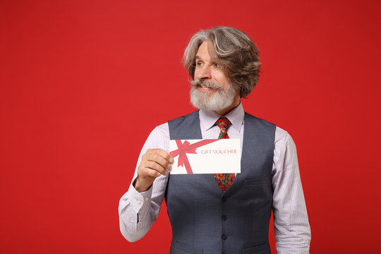 Handsome elderly gray-haired mustache bearded man in classic shirt vest and colorful tie isolated on red background. People lifestyle concept. Mock up copy space. Hold gift certificate, looking aside. - Powered by Adobe