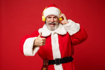 Fototapeta na wymiar Elderly gray-haired mustache bearded Santa man in Christmas hat posing isolated on red background. New Year 2020 celebration concept. Mock up copy space. Listen music with headphones showing thumb up.