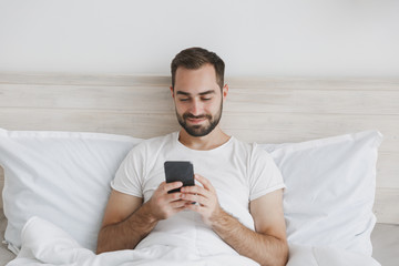 Calm young handsome bearded man lying in bed with white sheet pillow blanket in bedroom at home. Male spending time in room with mobile phone. Rest relax good mood lifestyle concept Mock up copy space