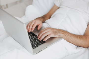 Close up cropped image of man lying in bed with white blanket in bedroom at home. Male spend time in room using pc laptop. Hands on keyboard. Rest relax good mood lifestyle concept. Mock up copy space