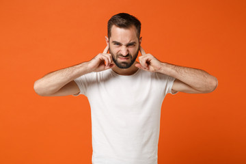 Naklejka premium Displeased young man in casual white t-shirt posing isolated on bright orange background studio portrait. People sincere emotions lifestyle concept. Mock up copy space. Covering ears with fingers.