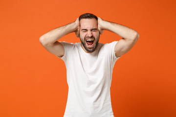Fototapeta na wymiar Frustrated young man in casual white t-shirt posing isolated on bright orange background in studio. People lifestyle concept. Mock up copy space. Screaming, keeping eyes closed, putting hands on head.
