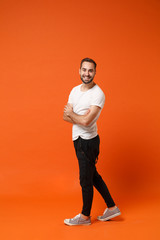 Fototapeta na wymiar Side view of smiling young man in casual white t-shirt posing isolated on orange wall background studio portrait. People lifestyle concept. Mock up copy space. Holding hands crossed, looking camera.
