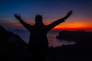 Man traveler silhouette hiking at sunset on Santorini island. Happy tourist raised arms feeling free, admiring landscape